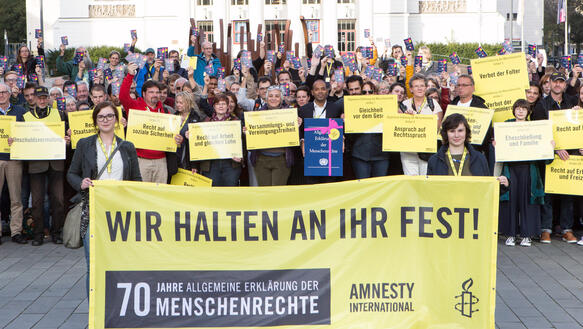 Eine Gruppe von Menschen hält gelbe Schilder hoch, auf denen jeweils ein Menschenrecht geschrieben steht. Im Vordergrund halten zwei Frauen ein großes Banner, auf dem "Wir halten an ihr fest! 70 Jahre Allgemeine Erklärung der Menschenrechte"